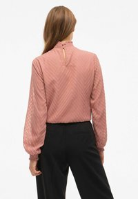 Różowa bluzka z długim rękawem z teksturowanym wzorem jodełki, wysokim falbaniastym kołnierzem oraz otworem na plecach, zestawiona z czarnymi spodniami.