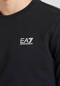 Sudadera negra con cuello redondo, que presenta un logo blanco de EA7 y el texto "EMPORIO ARMANI". Está hecha de algodón y tiene una textura suave.