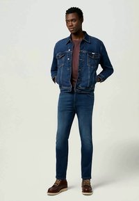 Jaqueta de ganga azul escura com dois bolsos no peito, usada sobre uma camisa bordô, emparelhada com jeans azul escuro e botas castanhas com atacadores.