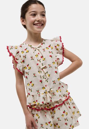 Fille souriante aux cheveux bruns porte un ensemble blouse et jupe crème avec un imprimé floral rouge et jaune et une bordure rouge sur les manches froncées et l'ourlet.