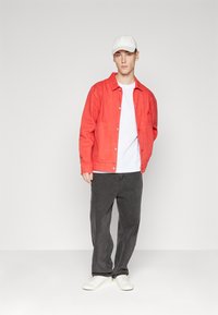 Sarkans džinsa jaka, balts T-krekls, melni taisni džinsi un balti sporta apavi; jaka ir aprīkota ar spiedpogām un krūšu kabatu.