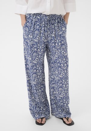 Osoba nosiaca voľné modré nohavice s bielymi kvetinovými vzormi, bielu košeľu, ruky v vreckách a čierne žabky, stojaca na bielom pozadí.