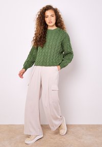 Grön kabelstickad tröja med långa ärmar, kombinerad med ljusbeige vida cargobyxor med två sidofickor. Sneakers är ljusbeige.