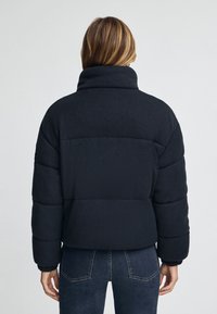 Mujer vestida con una chaqueta acolchada negra y un collar alto, y unos vaqueros oscuros, de pie de espaldas contra un fondo claro y liso.