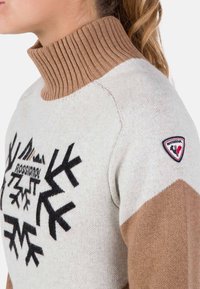 Maglione con collo alto a coste marrone, corpo crema e maniche marrone chiaro. Presenta motivi a fiocco di neve neri e una toppa con logo sulla spalla.