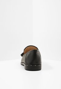 Chaussure en cuir noir avec un bout rond et un gland à l'arrière. Elle présente une texture lisse et des clous en argent décoratifs le long de la semelle.