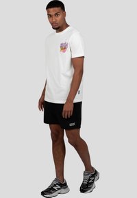 Bílá bavlněná tričko s barevným grafickým designem na hrudi, spárované s černými šortkami s logem. Šedé pozadí.