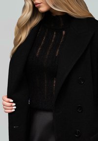 Donna bionda che indossa un maglione nero a maglia testurizzata e un cappotto nero doppiopetto, aperto con le unghie curate di bianco.