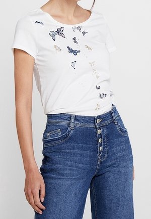 Kvinde iført hvid kortærmet skjorte med blå og beige sommerfugleprint og højttaljede blå jeans med knapper.