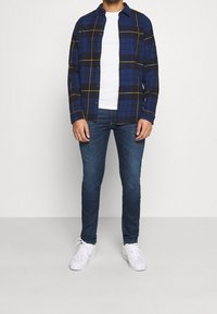 Camicia di flanella a scacchi blu e gialli, con una T-shirt bianca sotto, abbinata a jeans scuri e sneakers bianche. Presenta un bottone frontale e colletto standard.