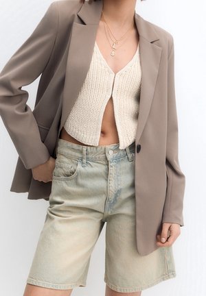 Personne portant un blazer beige par-dessus un gilet court en maille crème et un short bermuda en denim délavé clair, accessoirisé avec des colliers dorés superposés.