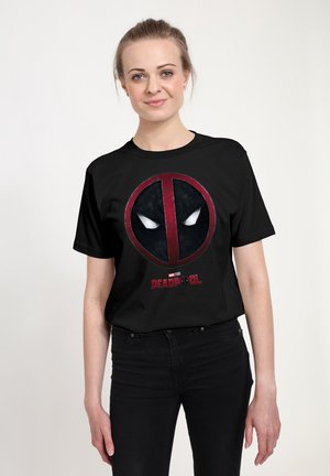 Kvinde med lys hud og blondt hår bundet tilbage, iført en sort Deadpool T-shirt og sorte bukser mod en ensfarvet hvid baggrund.
