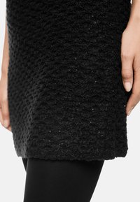 Schwarzes, strukturiertes Strickkleid mit einem erhabenen Muster und dezentem Schimmer, kombiniert mit schwarzen Leggings, zeigt eine Nahaufnahme des Saums und des Materials.