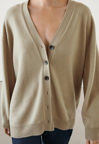 Beige oversized cardigan with a V-neck, large buttons, ribbed hems, and a soft texture. Worn over dark jeans, featuring a relaxed fit.
