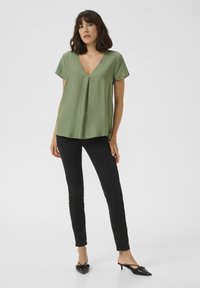 Blouse olive vert à manches courtes avec un décolleté en V, coupe ample et texture subtile, associée à un pantalon skinny noir et des escarpins noirs à bouts pointus.