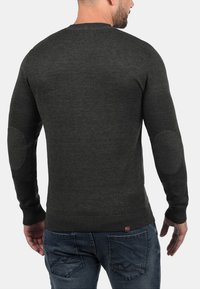 Pull en maille gris foncé avec un col rond, des poignets côtelés et des coudières. Porté avec un jean en denim bleu.