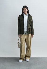 Olijfgroene jas met twee klepzakken, witte zip-up trui, gestreept shirt, beige wijde broekspijpen en witte sneakers. Kleine handtas.