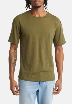 Uomo che indossa una t-shirt a maniche corte verde oliva con un piccolo logo sul petto, jeans azzurri e tatuaggio visibile sull'avambraccio sinistro.