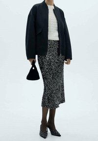 Czarna kurtka bomberka, kremowy sweter o fakturze dzianiny, srebrna spódnica z cekinami, czarna torebka oraz czarne szpilki z białymi ćwiekami na noskach.