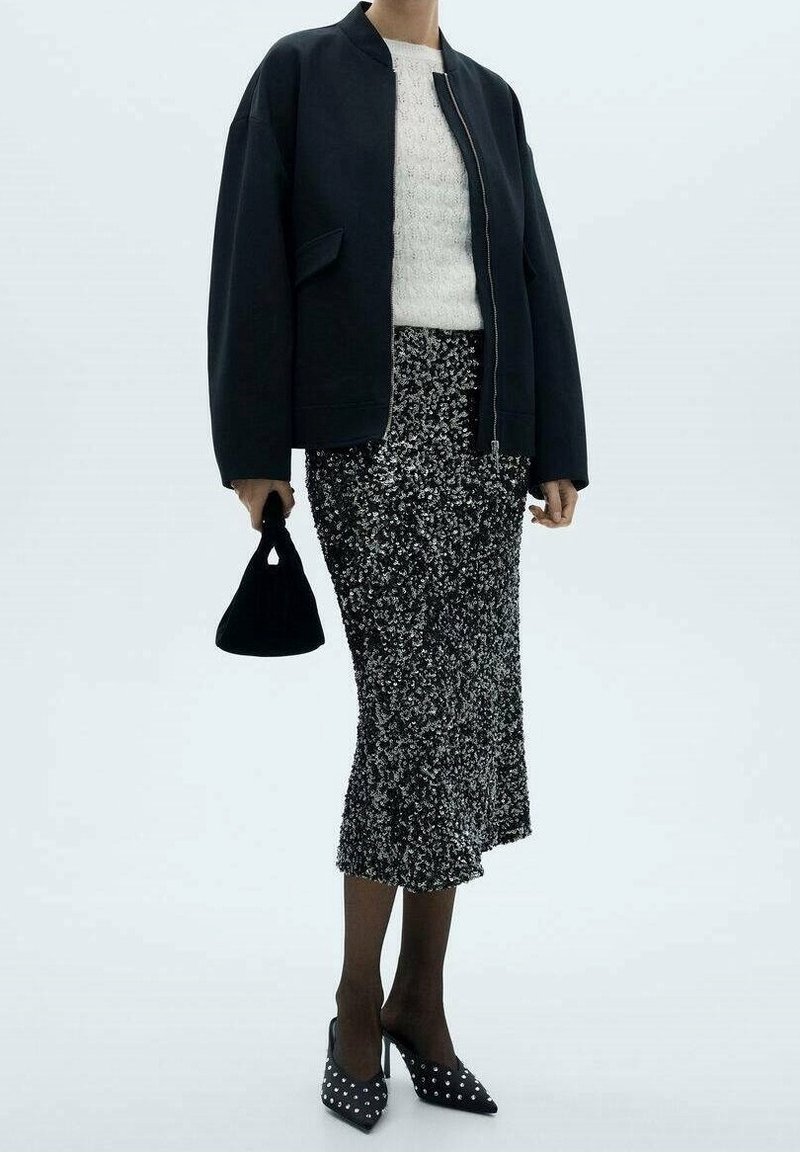 Czarna kurtka bomberka, kremowy sweter o fakturze dzianiny, srebrna spódnica z cekinami, czarna torebka oraz czarne szpilki z białymi ćwiekami na noskach.