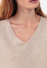 Pull à col en V de couleur beige clair. Confectionné en tissu doux et texturé avec un motif de tricot visible. Présente des détails de couture subtils le long de l'encolure.