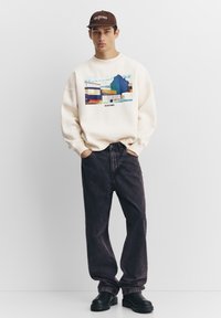 Crèmesweatshirt met een kleurrijk grafisch ontwerp aan de voorkant, gecombineerd met donkerblauwe spijkerbroek en zwarte chunky laarzen, en een bruine pet.