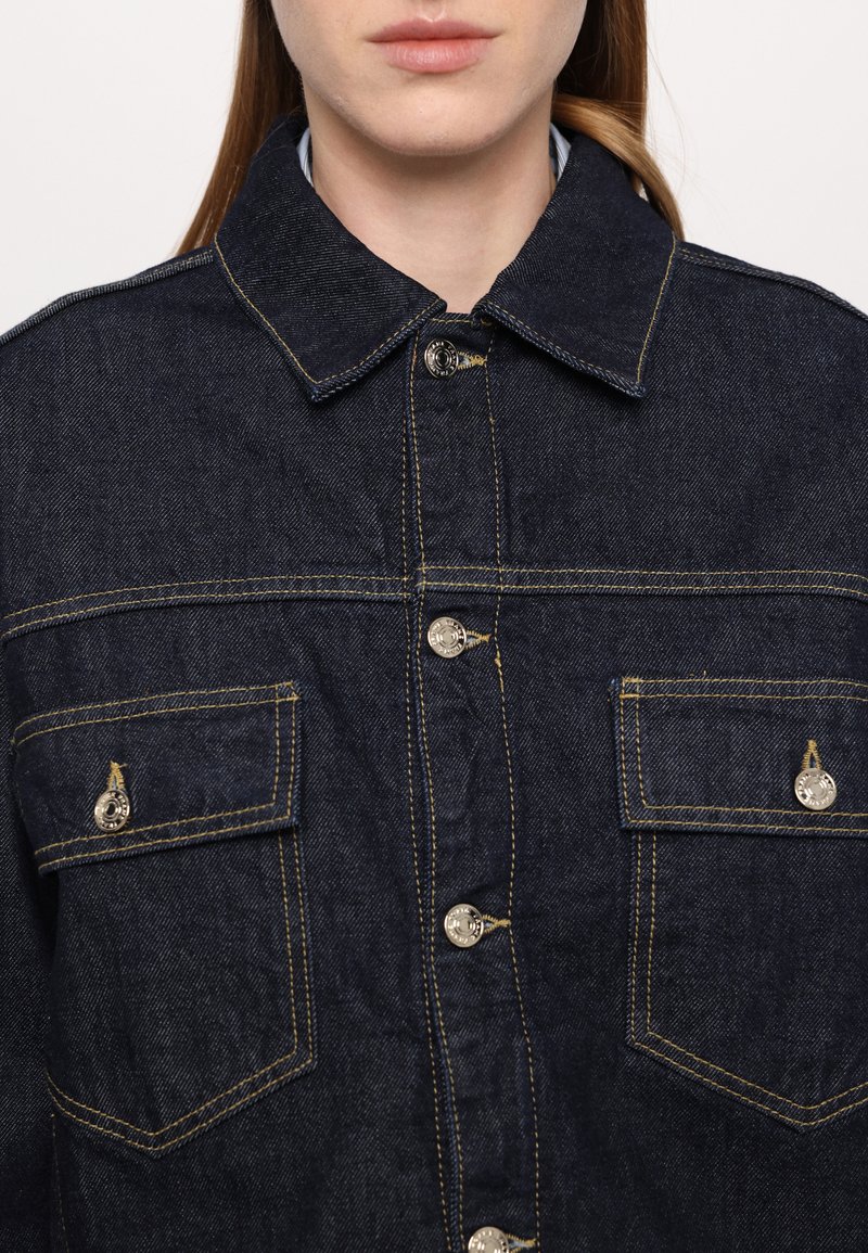 Giacca in denim blu scuro con bottoni davanti, due tasche sul petto e cuciture gialle a contrasto. Design classico con colletto.