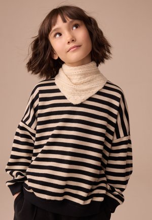 Junge Person mit kurzen braunen Haaren, die einen beigen gestrickten Halswärmer und einen schwarz-beigen gestreiften Pullover trägt und nach oben schaut.