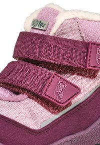 Scarpe di stoffa rosa con cinturini in velcro, superfici testurizzate e un logo argento. Presentano una fodera morbida e un emblemma a forma di orsetto sul cinturino.