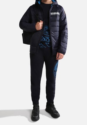 Bambino con giacca puffer blu navy, felpa con grafica, pantaloni jogger abbinati, sneaker nere e una borsa nera a tracolla su una spalla.
