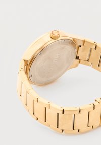 Χρυσό χρώμα μεταλλικό ρολόι χειρός με συνδεδεμένο λουρί, στρογγυλό πίσω καπάκι με χαραγμένο το όνομα της μάρκας και κορώνα που φέρει τη σημείωση "HUGO" με κόκκινο χρώμα.