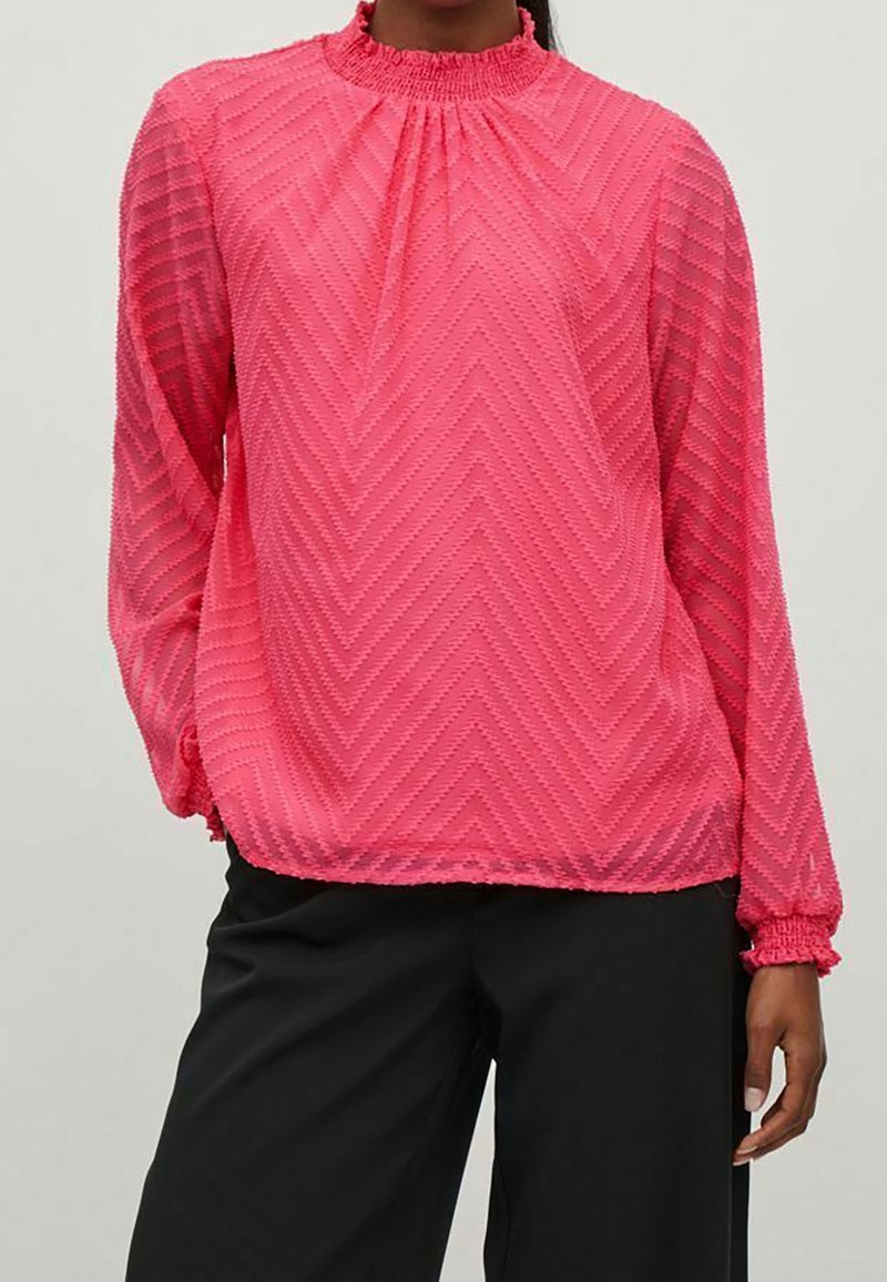 Blusa rosa de manga larga con un cuello alto fruncido, que presenta un patrón de zigzag y puños elásticos, combinada con pantalones negros.