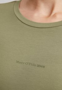 Olīvu zaļš pieguļošs T-krekls ar noapaļotu apkakli un īsām piedurknēm, ar izsmalcinātu iegravētu logotipu "Marc O'Polo DENIM" netālu no apkakles.