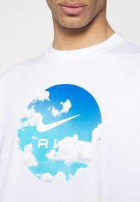 Bílá triko s kulatou modrou grafikou, na které jsou mraky a logo Nike nad slovem "AIR" v tučných bílých písmenech.