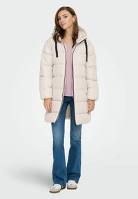 Puffjacke in hellbeige mit Reißverschluss vorne, Kapuze und schwarzen Kordeln. Getragen über einem hellrosa Oberteil und kombiniert mit blauen Schlagjeans.