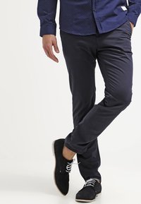 Navy Slim-Fit-Hosen aus einem glatten Stoff, kombiniert mit schwarzen Wildlederschuhen mit weißen Schnürsenkeln und einer hellen Sohle.