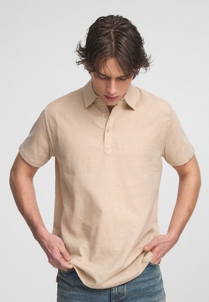 Polo beige a maniche corte con colletto, chiusura con tre bottoni e finitura testurizzata, abbinato a jeans in denim blu.