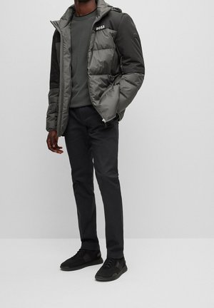 Mężczyzna w szarej ocieplanej kurtce z logo „BOSS”, ciemnej bluzie, czarnych spodniach i czarnych trampkach, stojący na tle jasnego, jednolitego tła.