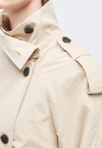 Κοντινό πλάνο μπεζ καμπαρντίνας με μαύρα κουμπιά και επιγονατίδες στους ώμους, με ψηλό γιακά, που φοριέται από άτομο.