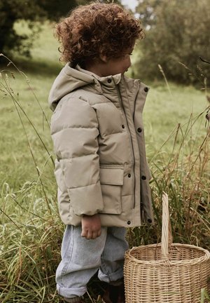 Enfant aux cheveux bouclés portant une doudoune beige et un jean clair, se tient à l'extérieur dans l'herbe à côté d'un panier en osier.