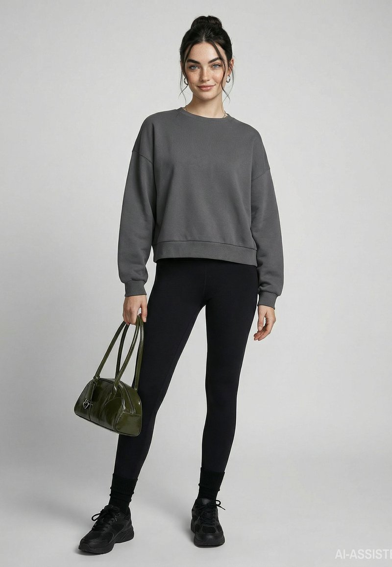 Jeune femme aux cheveux foncés en chignon, portant un sweat-shirt gris, des leggings noirs, des baskets noires, et tenant un sac à main vert.