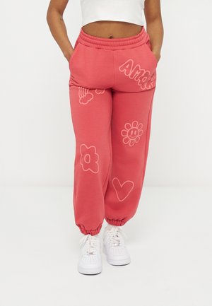 Γυναίκα που φοράει κοραλλί παντελόνι φόρμας με ελαστικά μανσέτες και σχεδιασμούς ενός λουλουδιού, καρδιάς, ουράνιου τόξου και το κείμενο "Amar", συνδυασμένο με λευκά αθλητικά παπούτσια.