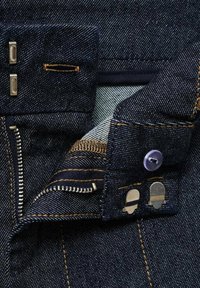 Mörkblå denimtyg med en synlig dragkedja, metallbeslag och kontrasterande orange sömmar. Har en lila knapp och foderdetalj.