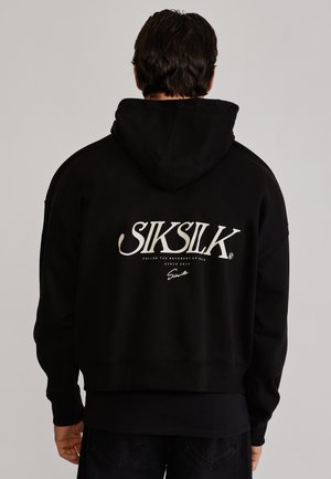 Homme portant un sweat à capuche noir avec le logo texte "SIKSILK" et un slogan plus petit imprimés au dos, debout devant un fond gris clair.
