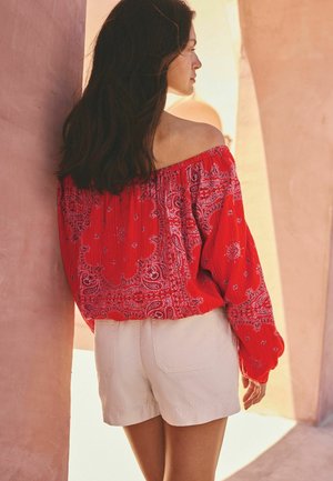 Woman with long dark hair wearing a red off-shoulder patterned blouse and white shorts, standing near a peach-colored wall with arches.