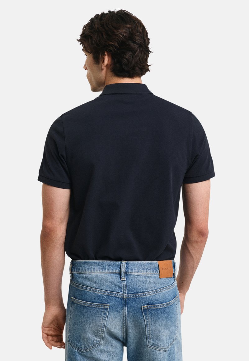 Dunkelblaues Poloshirt mit kurzen Ärmeln und klassischem Kragen, kombiniert mit hellblauen Jeans aus Denim, die einen braunen Lederpatch am Bund haben.