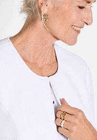 Witte gestructureerde jas met een ronde hals, vastgemaakt met een enkele knop. Accessoires zijn gouden hoopsieraden en gelaagde ringen.