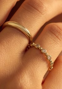 Bague de mariage en or avec une surface lisse et polie, accompagnée d'une délicate bague en or ornée d'accents de pierres circulaires. Peau texturée visible.