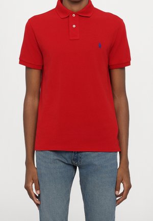 Man wearing red short-sleeve polo shirt with blue embroidered logo on chest and light blue jeans against white background.