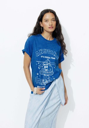 T-shirt en coton bleu arborant un personnage de dessin animé sur un skateboard avec le texte "TOUJOURS L'HEURE DE LA PIZZA." Col rond et manches retroussées.